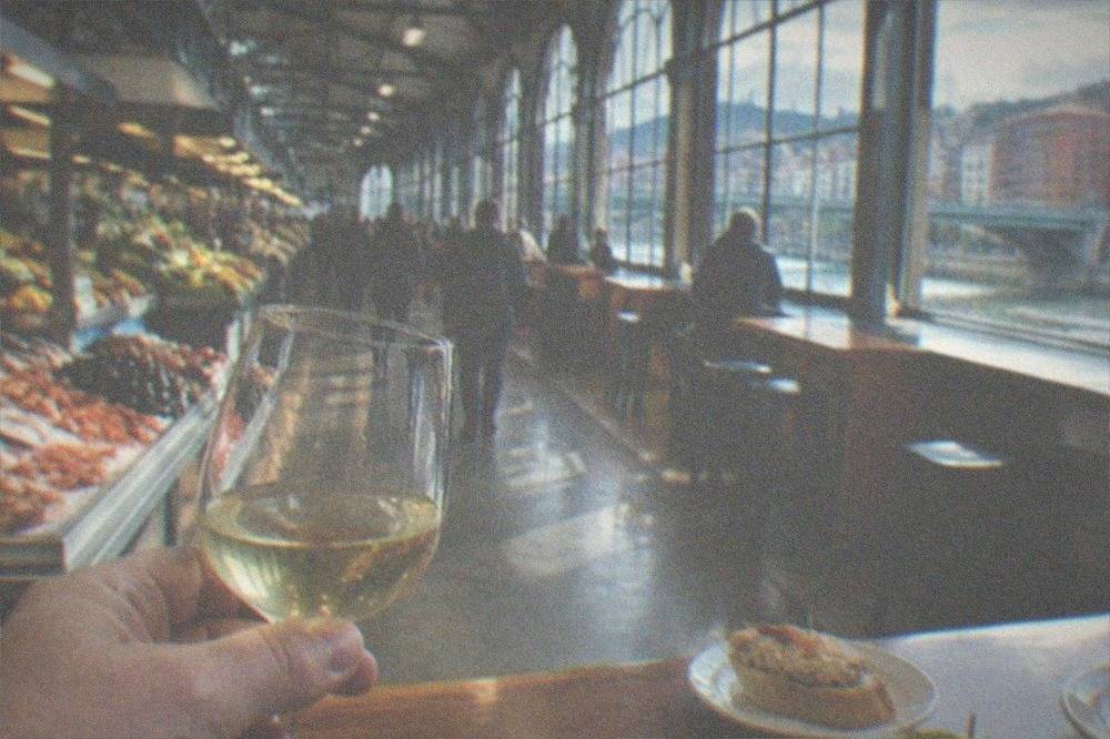 A hand holding a small glass of txakoli white wine above a wooden bar top with pintxo plates