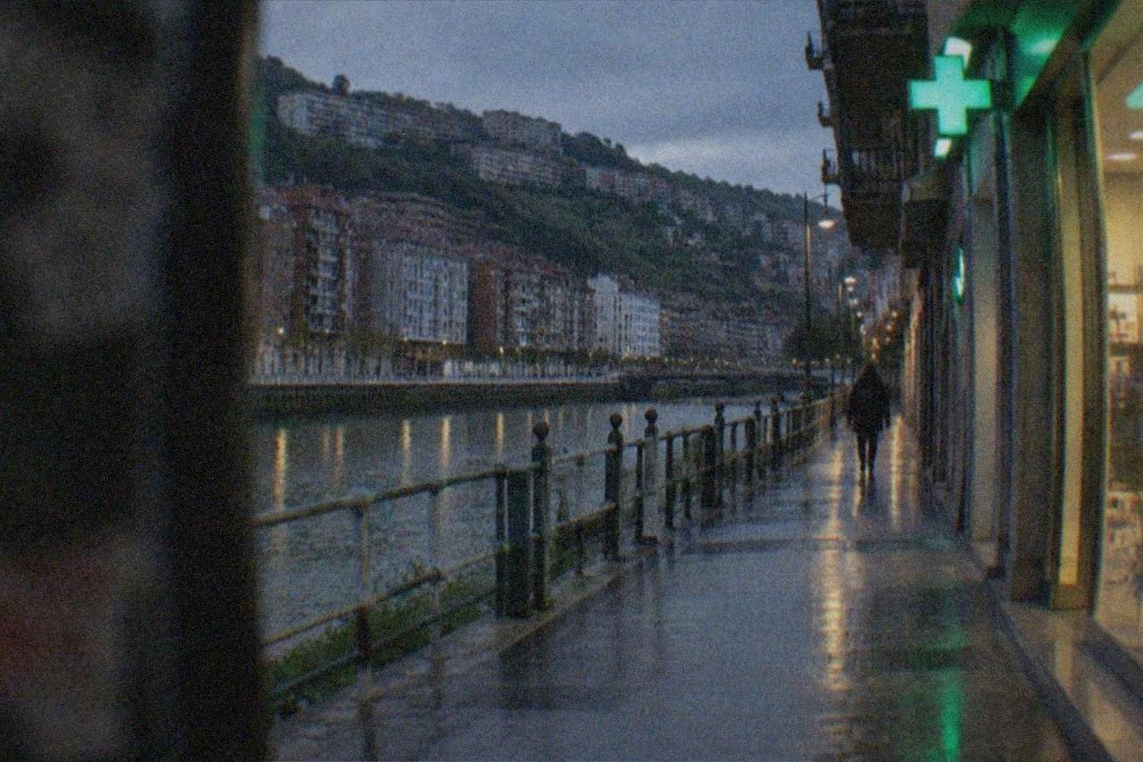 A pharmacy storefront on a Bilbao street with a lit green cross sign above the door