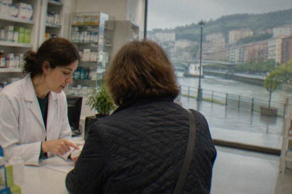 A pharmacist in a white coat advising a customer at a pharmacy counter in Bilbao