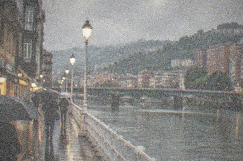 A rainy evening on a Bilbao street with pedestrians under umbrellas near lit shopfronts