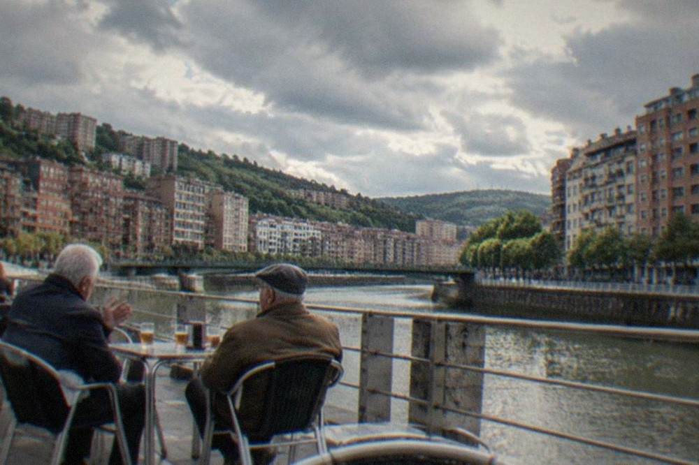 Two older men talking at an outdoor terrace table in a Bilbao square