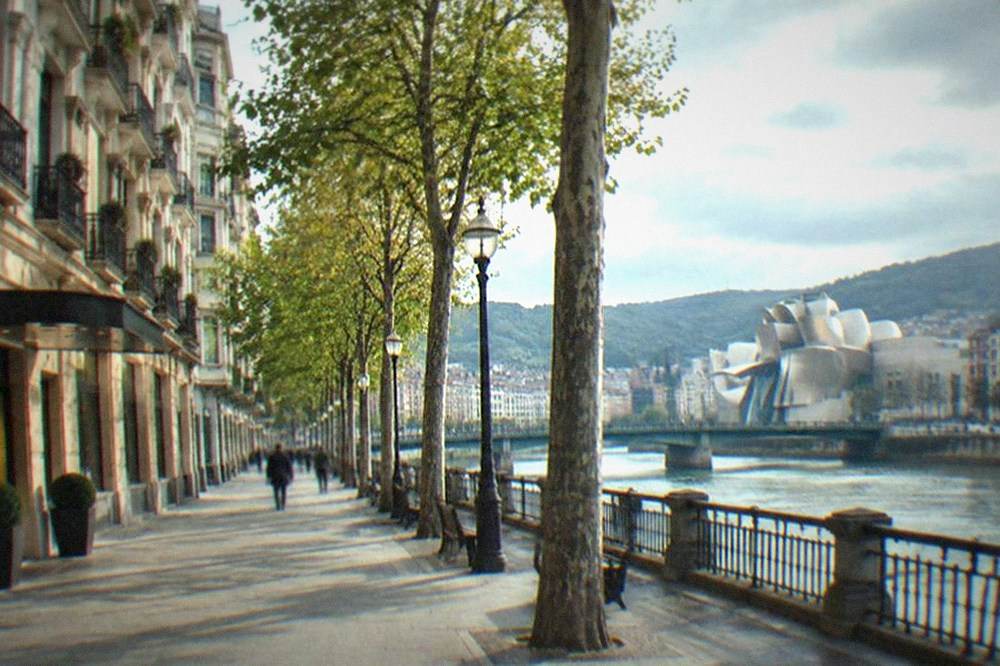 Facade of a hotel on a tree-lined street in Bilbao's Ensanche district with the Guggenheim museum visible in the distance