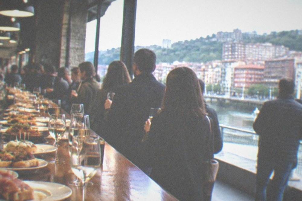 A pintxos bar counter in Bilbao crowded with small plates, glasses of txakoli and standing customers