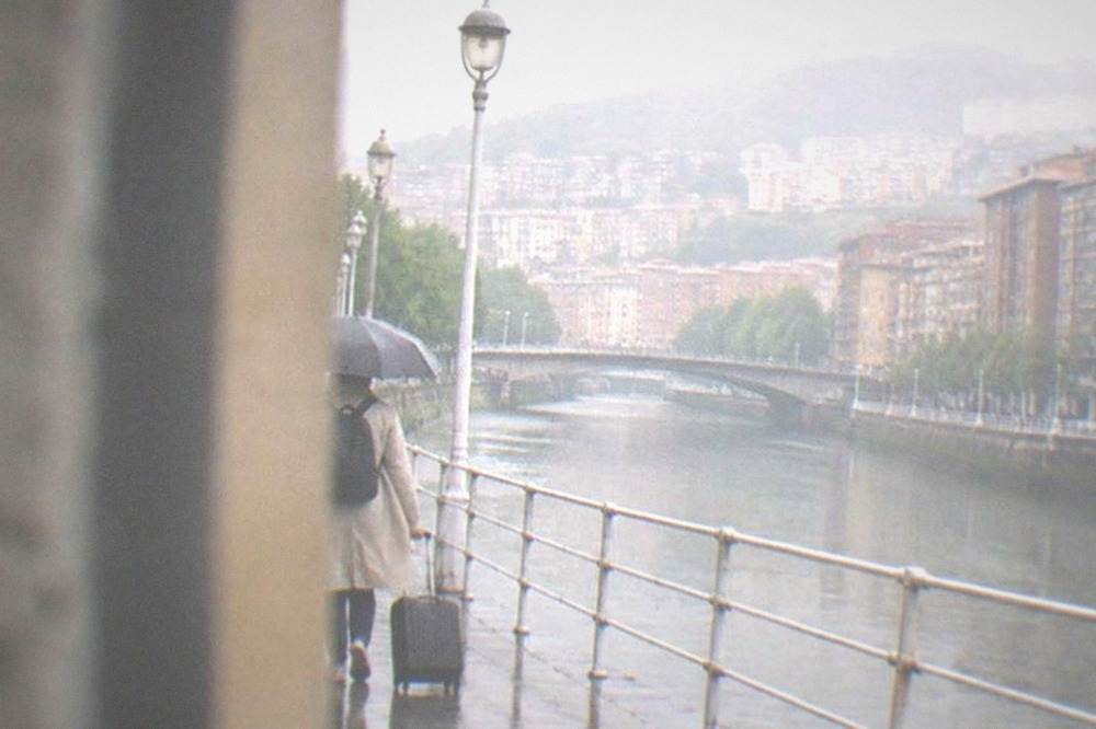 A traveler with a small carry-on walking under an umbrella along a wet Bilbao street