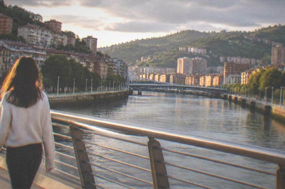 A traveler walking bag-free across a Bilbao bridge at golden hour with the estuary behind