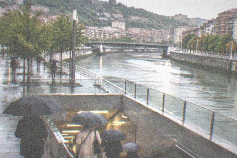 Commuters with umbrellas entering a metro station entrance on a rainy Bilbao evening
