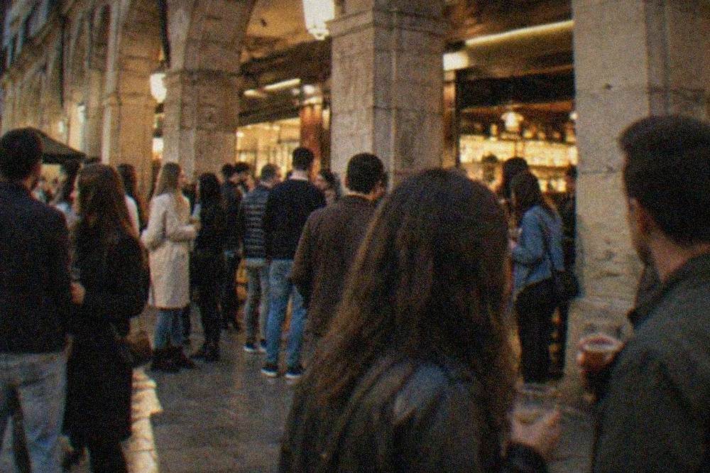 Crowded square in Bilbao old town at night with people holding drinks outside lit bar fronts under stone arcades