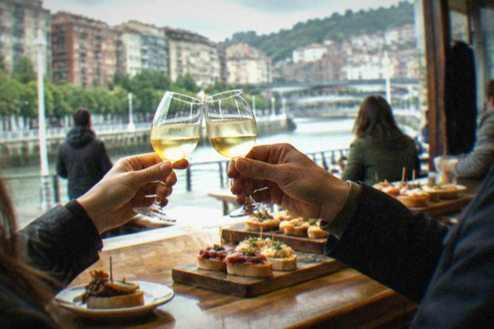 Two people clinking small glasses of txakoli wine at a busy wooden pintxos bar counter in Bilbao