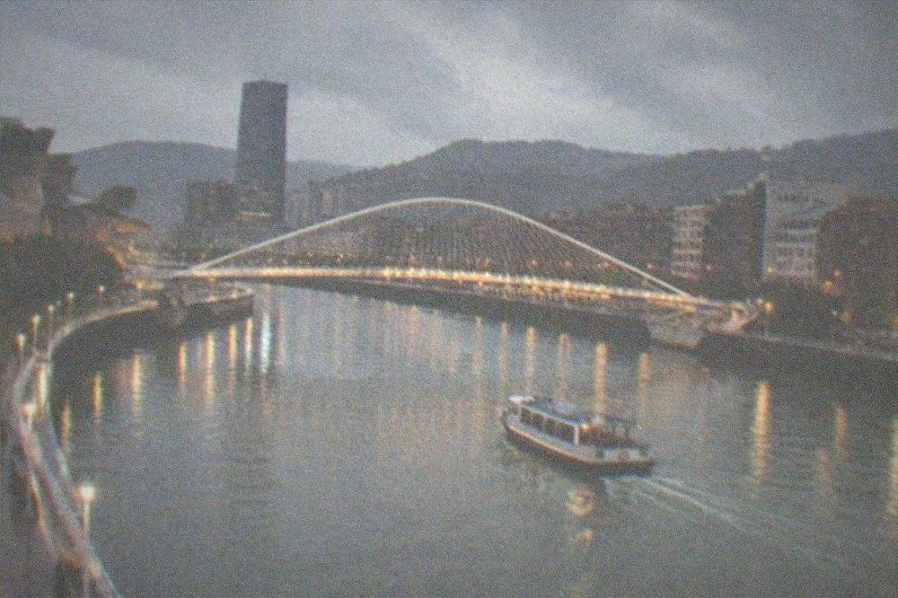 Illuminated Zubizuri footbridge over the Nervion river in Bilbao at dusk with city lights reflecting on the water