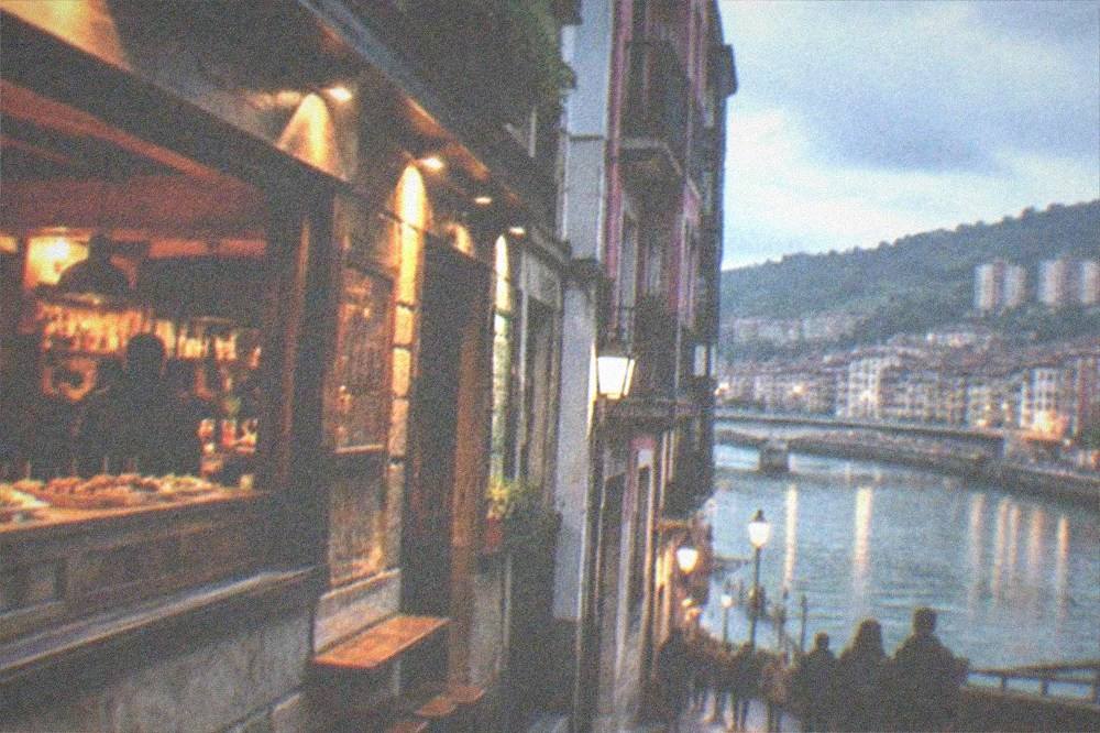 Lit facade of a traditional Bilbao pintxos bar in the evening with warm light spilling onto a narrow old town street