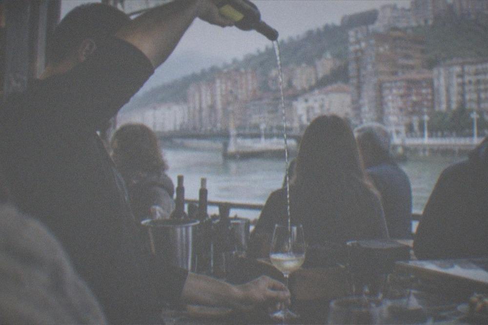 Bartender pouring txakoli white wine from a height into a glass at a crowded Bilbao bar counter