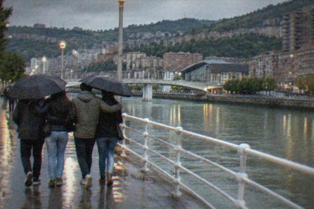 Group of friends walking arm in arm along a lit Bilbao street at night wearing jackets and holding umbrellas