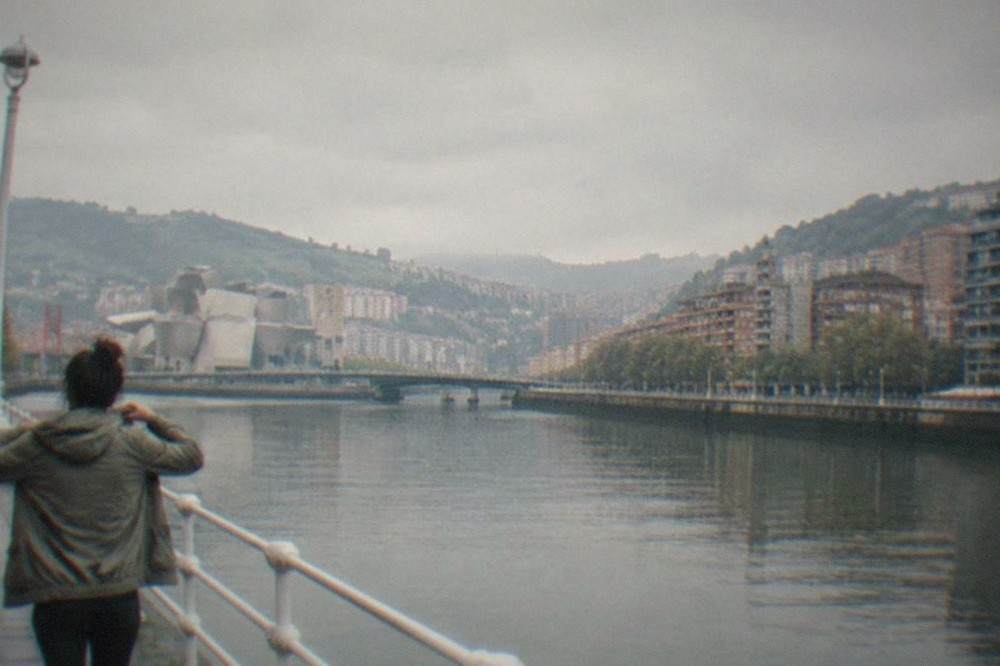 A traveler adjusting a light jacket beside the Bilbao estuary on an overcast day
