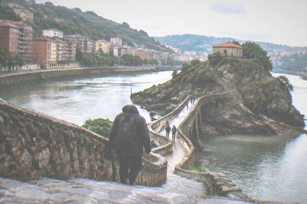 A hiker in a rain jacket walking the stone steps toward the Gaztelugatxe hermitage
