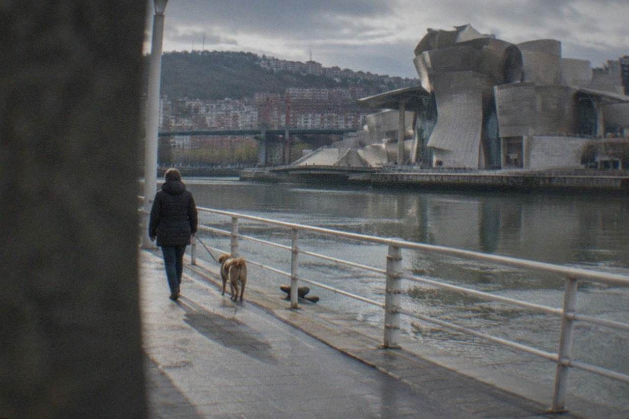 A dog on a lead beside a walker on a riverside promenade with modern museum architecture across the water