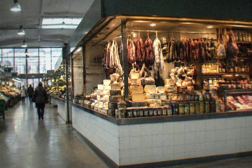 A market stall displaying Basque cheeses, charcuterie and tinned goods under warm lighting