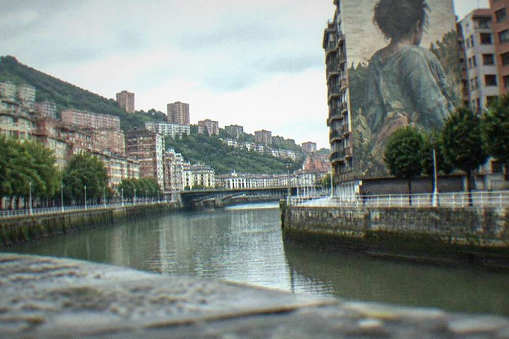 A tall gable wall in Bilbao covered by a large figurative mural seen from a narrow street