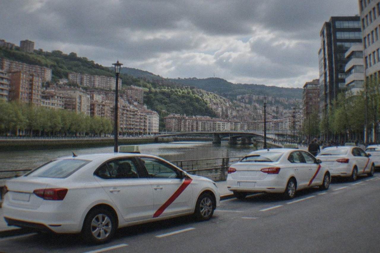 A line of white taxis with a red stripe parked at a rank on a Bilbao street with modern buildings behind