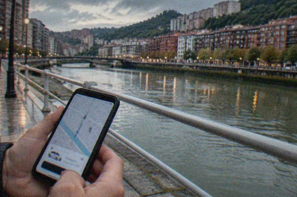 A person holding a smartphone showing a ride-booking app on a Bilbao sidewalk at dusk