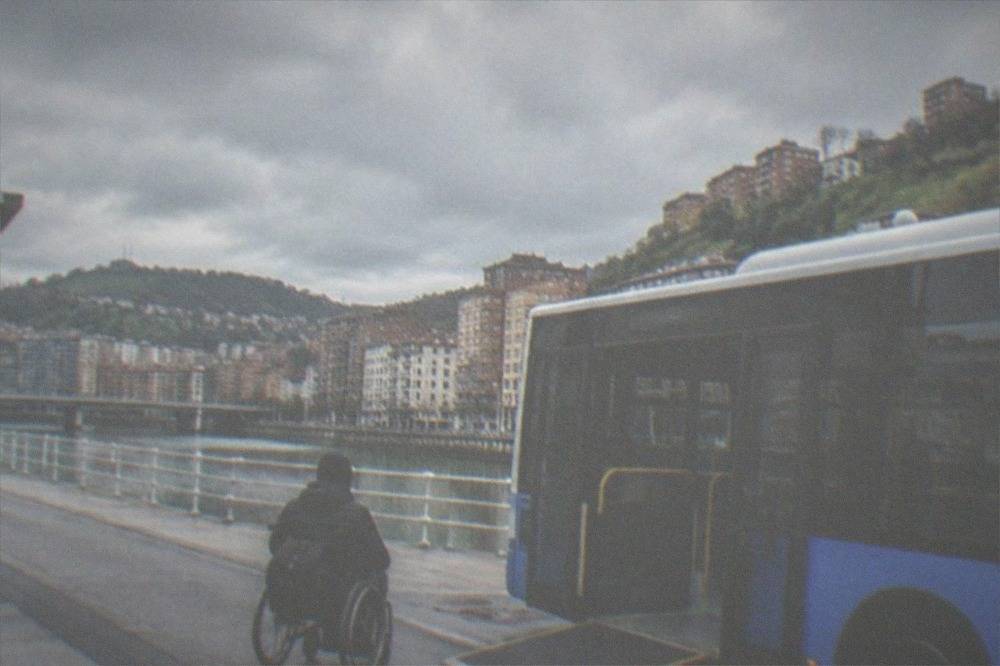 A low-floor city bus with a ramp lowering at a stop while a passenger with a wheelchair waits