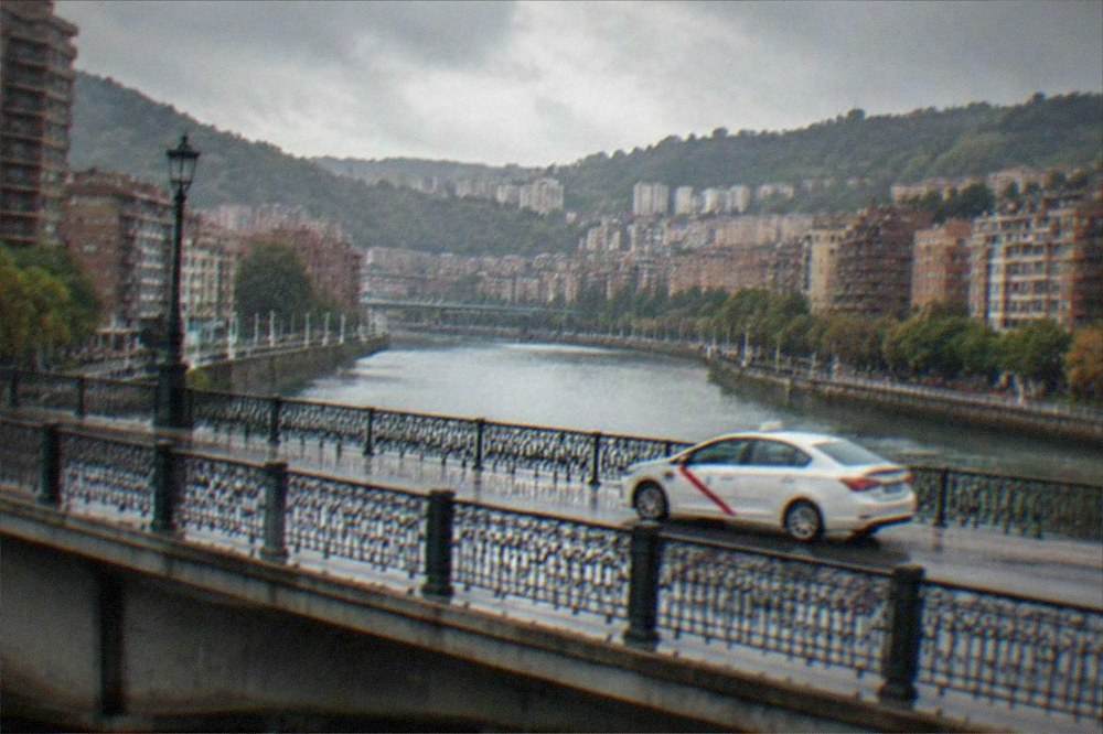 A white taxi with a red stripe driving across a bridge over the Bilbao estuary in daylight