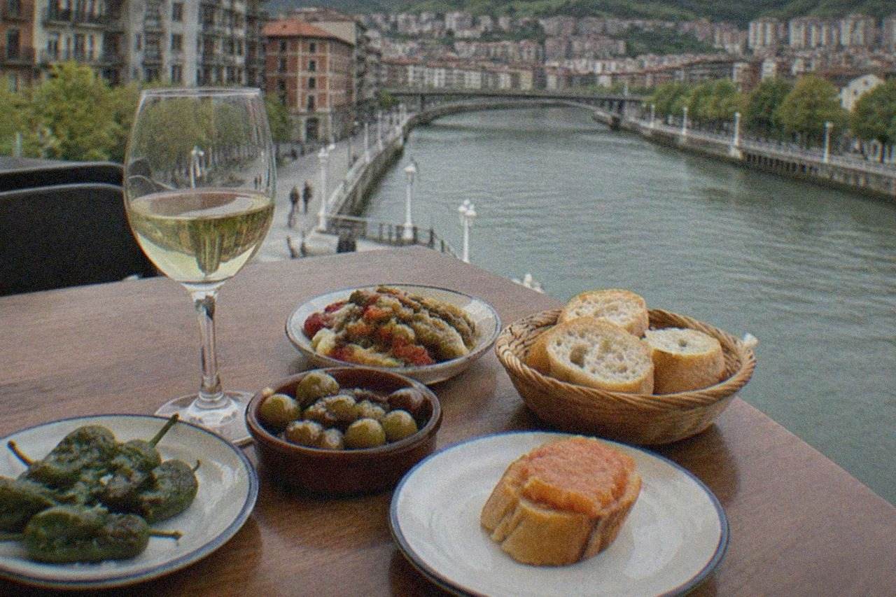 A table at a Bilbao pintxos bar set with small plates of vegetables, olives and bread, a glass of white wine beside them