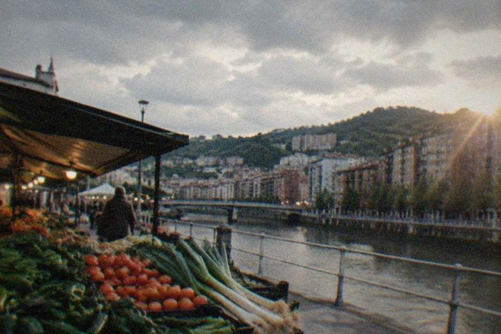 A Bilbao market stall piled with fresh green peppers, tomatoes and leeks under warm lights