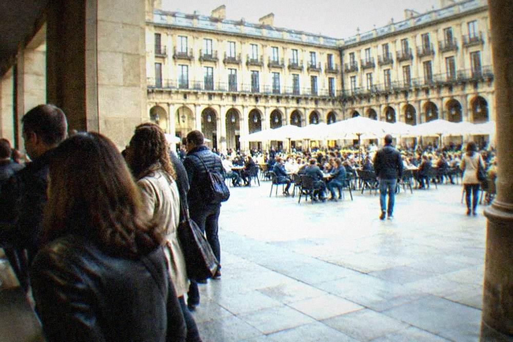 Crowds standing at a busy pintxos counter in Bilbao's Plaza Nueva, small plates on the bar top