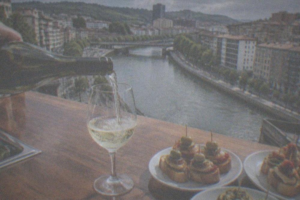 A glass of pale txakoli wine being poured at a bar counter with small vegetable pintxos waiting