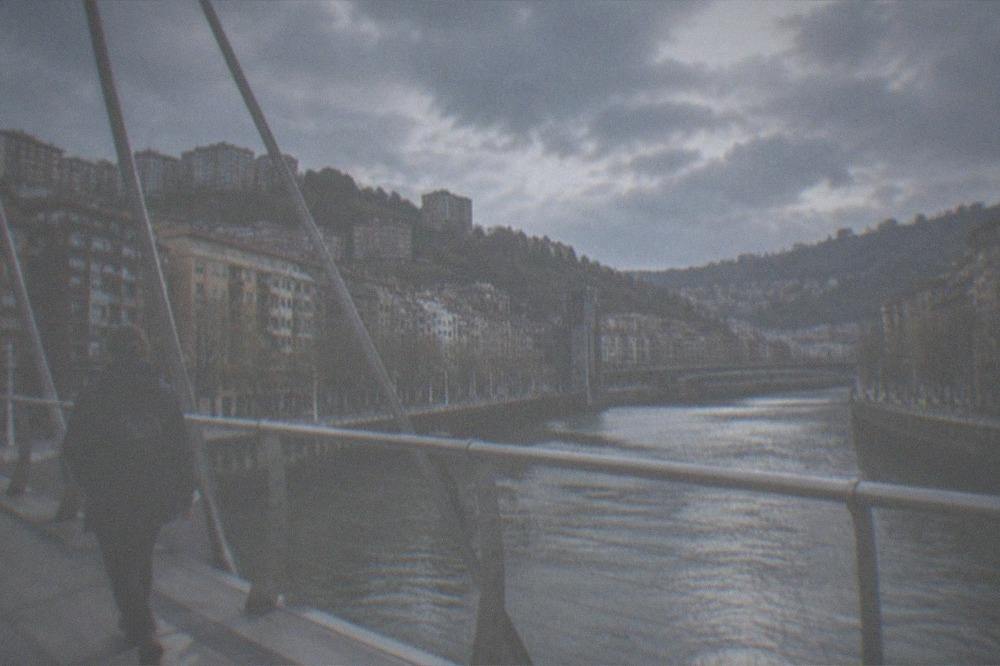 A traveller with a small backpack and rain jacket crossing a Bilbao bridge
