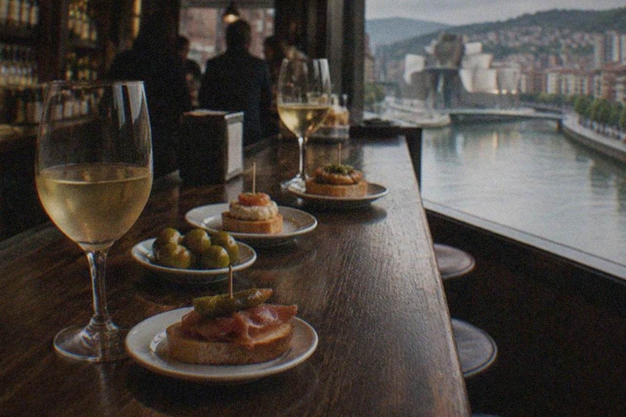 A dimly lit wine bar counter in Bilbao with glasses of white txakoli and small pintxo plates arranged on dark wood