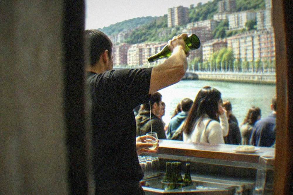 A bartender pouring txakoli from height into a small glass at a crowded Bilbao bar