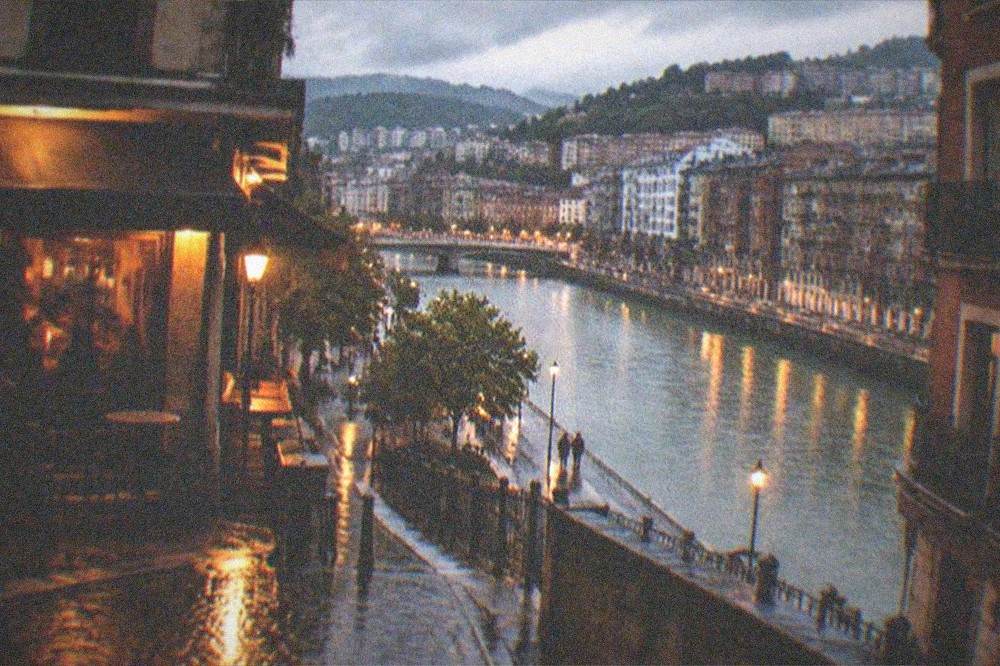 A Bilbao street at night with warm light spilling from a corner wine bar onto wet cobblestones