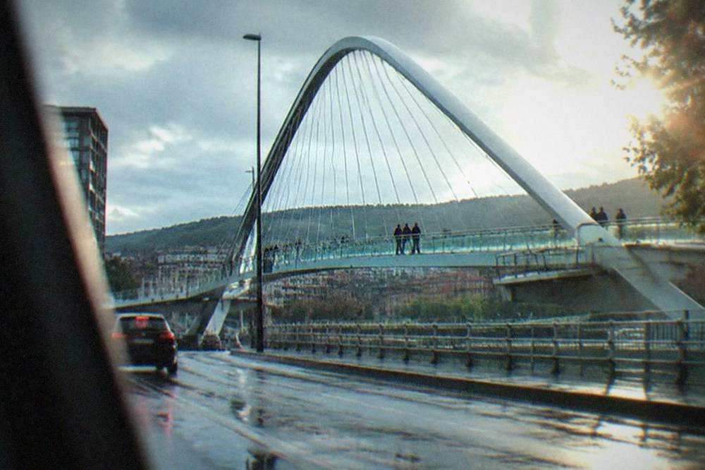A driver's view along a wet Bilbao street with the Zubizuri bridge ahead