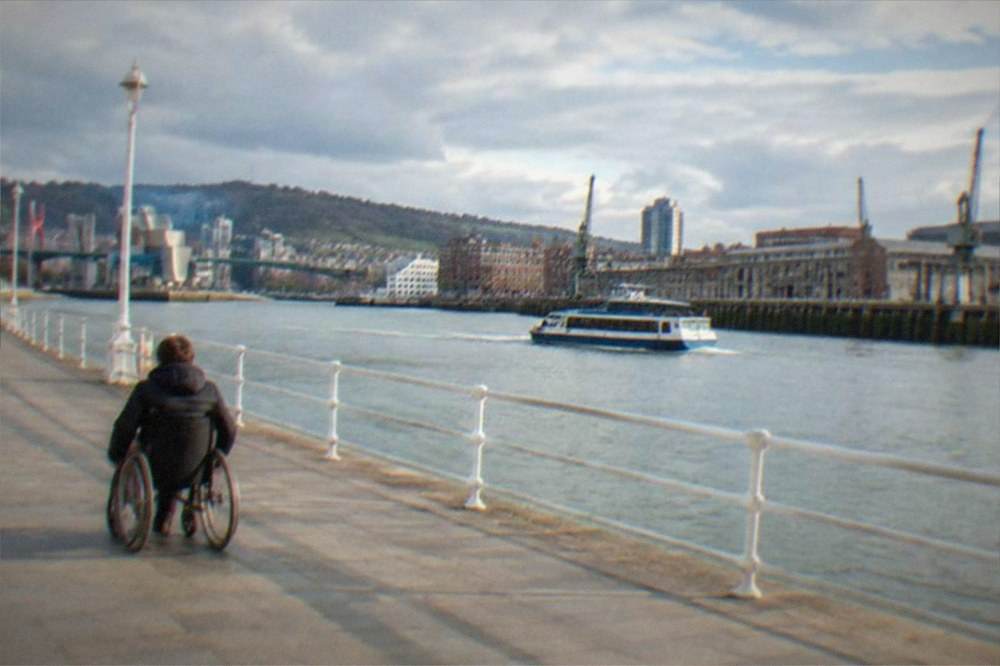 A wheelchair user rolling along the wide riverside promenade beside the Nervion estuary in Bilbao on a bright day