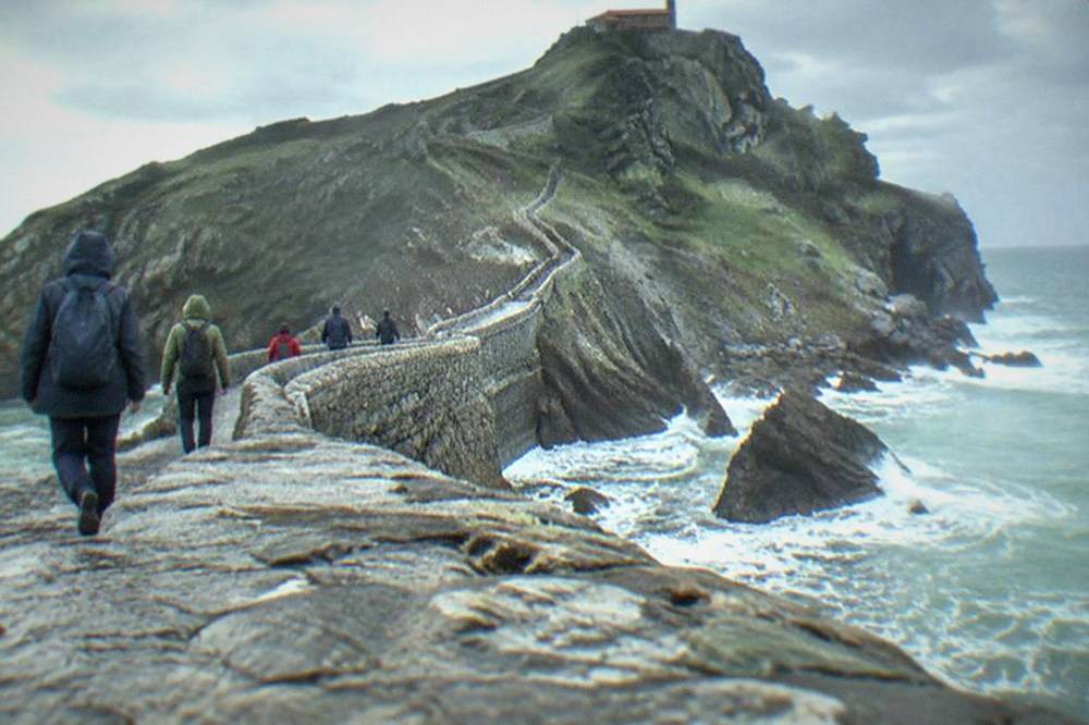 Walkers in rain jackets crossing a stone causeway over rough sea toward a rocky island in the Basque Country