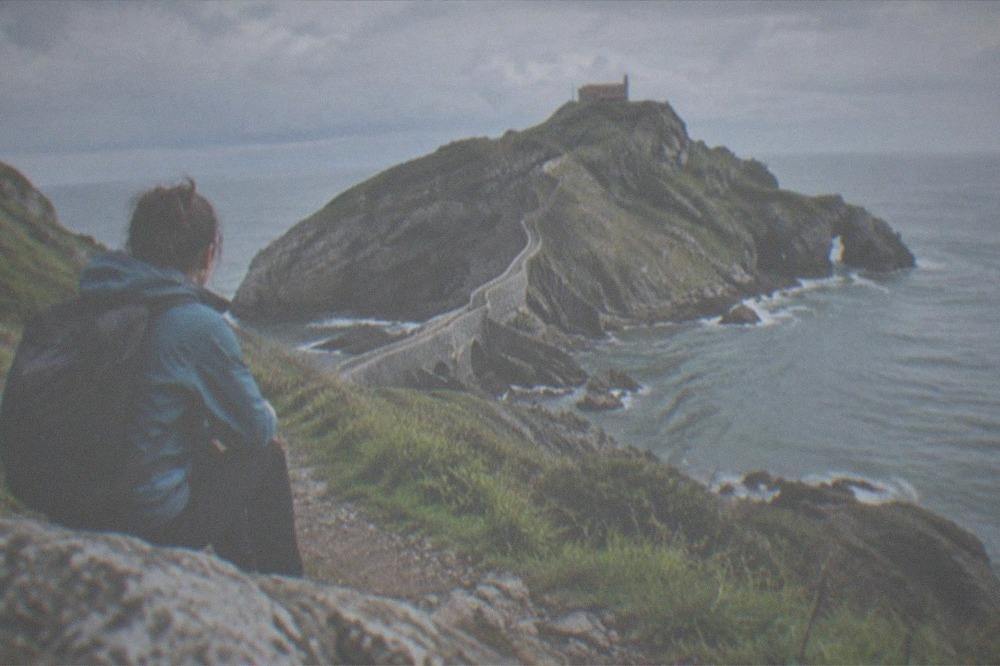 A hiker with a backpack and windbreaker resting on a coastal path above the sea in the Basque Country