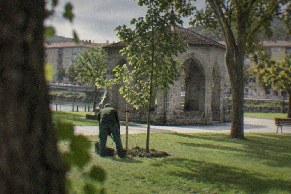 A gardener tending a young oak tree on a lawn beside a stone chapel-like pavilion in Gernika