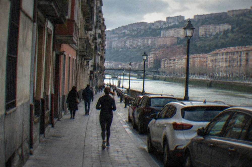 A narrow street in the Bilbao Old Town with cars parked along the edge and pedestrians walking