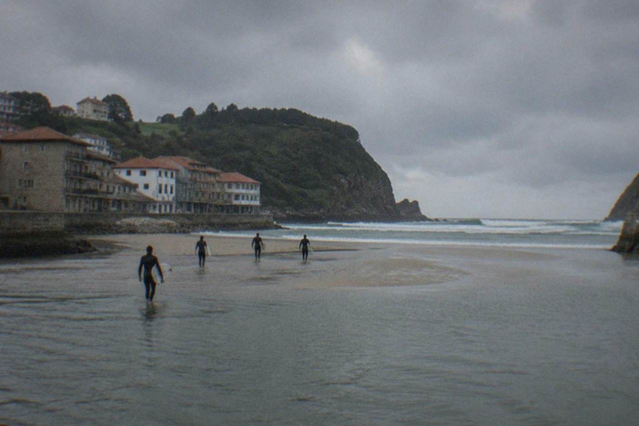 Surfers walking across a tidal sandbar toward rolling waves at a river mouth surrounded by green cliffs