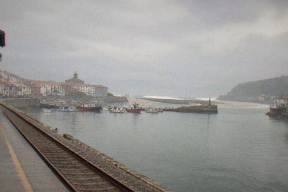 A single-track railway platform beside a small Basque coastal town with a harbour in the background