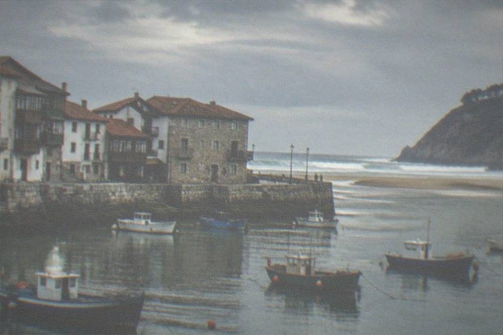 A small Basque fishing harbour with wooden boats and stone houses under an overcast sky