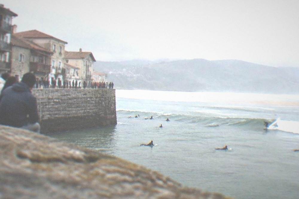 Onlookers lined along a stone harbour wall watching surfers ride waves below under grey skies