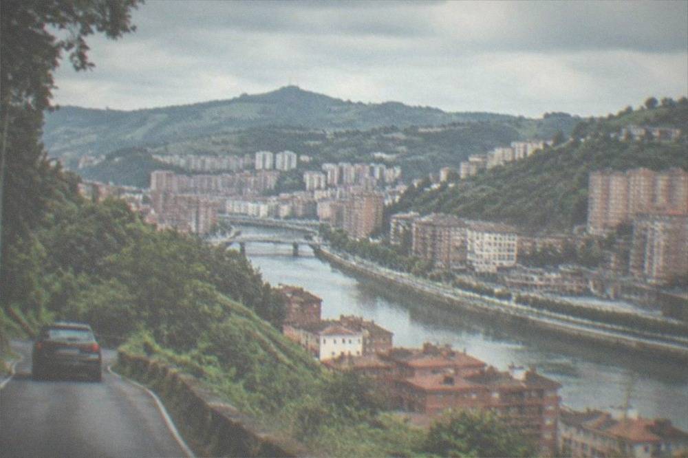 A car winding down a narrow green hillside road above the Basque coastline
