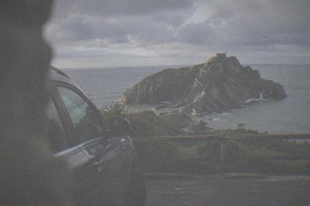A car parked at a coastal overlook facing the rocky islet and causeway of Gaztelugatxe