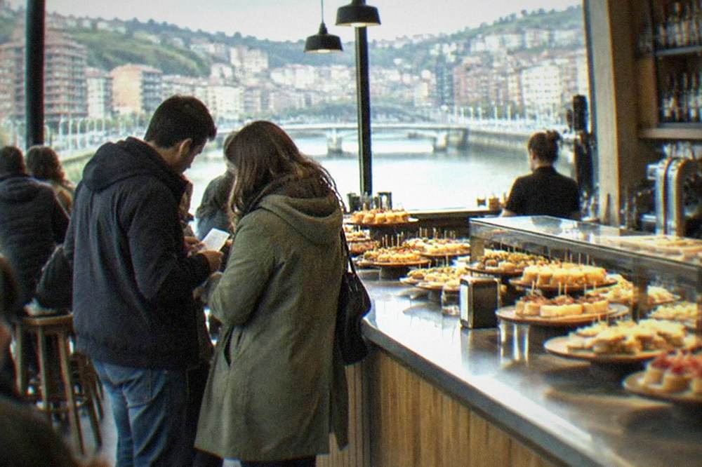 Two people comparing a bill at the counter of a busy pintxos bar in Bilbao