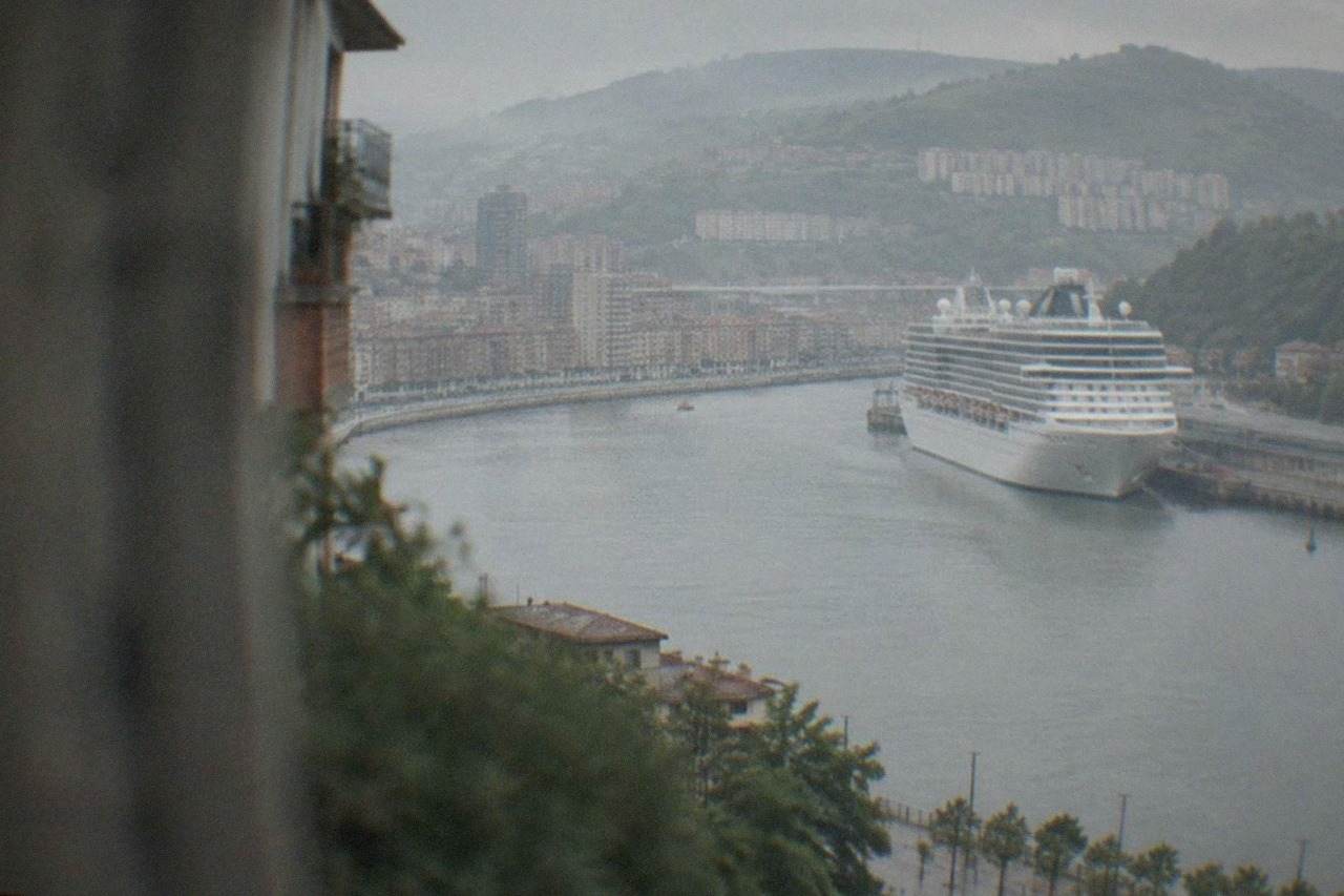 A large cruise ship docked at a terminal on the Bilbao estuary with green hills behind it