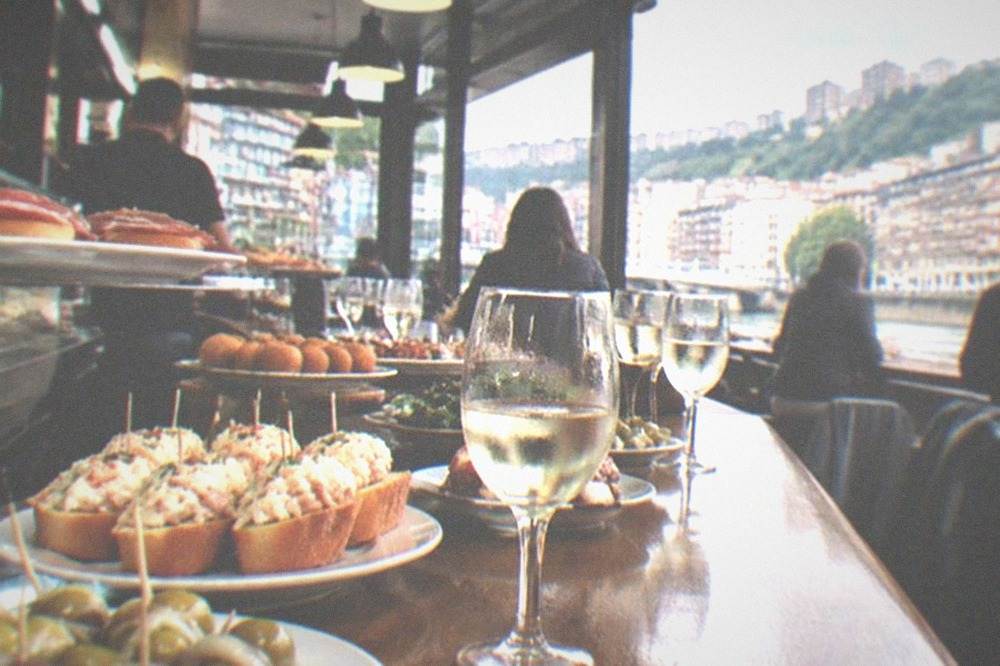 A pintxos bar counter in Bilbao crowded with small plates, gildas and txakoli glasses