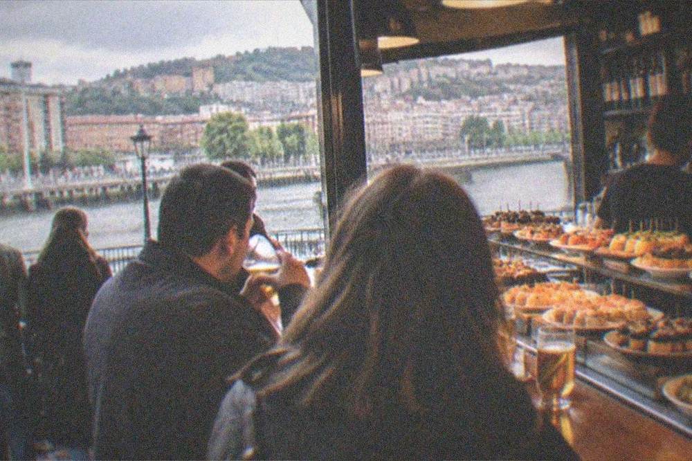 An evening crowd with drinks standing at the counter of a lit pintxos bar in Bilbao