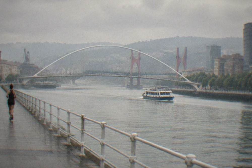 A white calatrava-style footbridge arching over the Nervion estuary with the red pylons of the La Salve bridge behind it